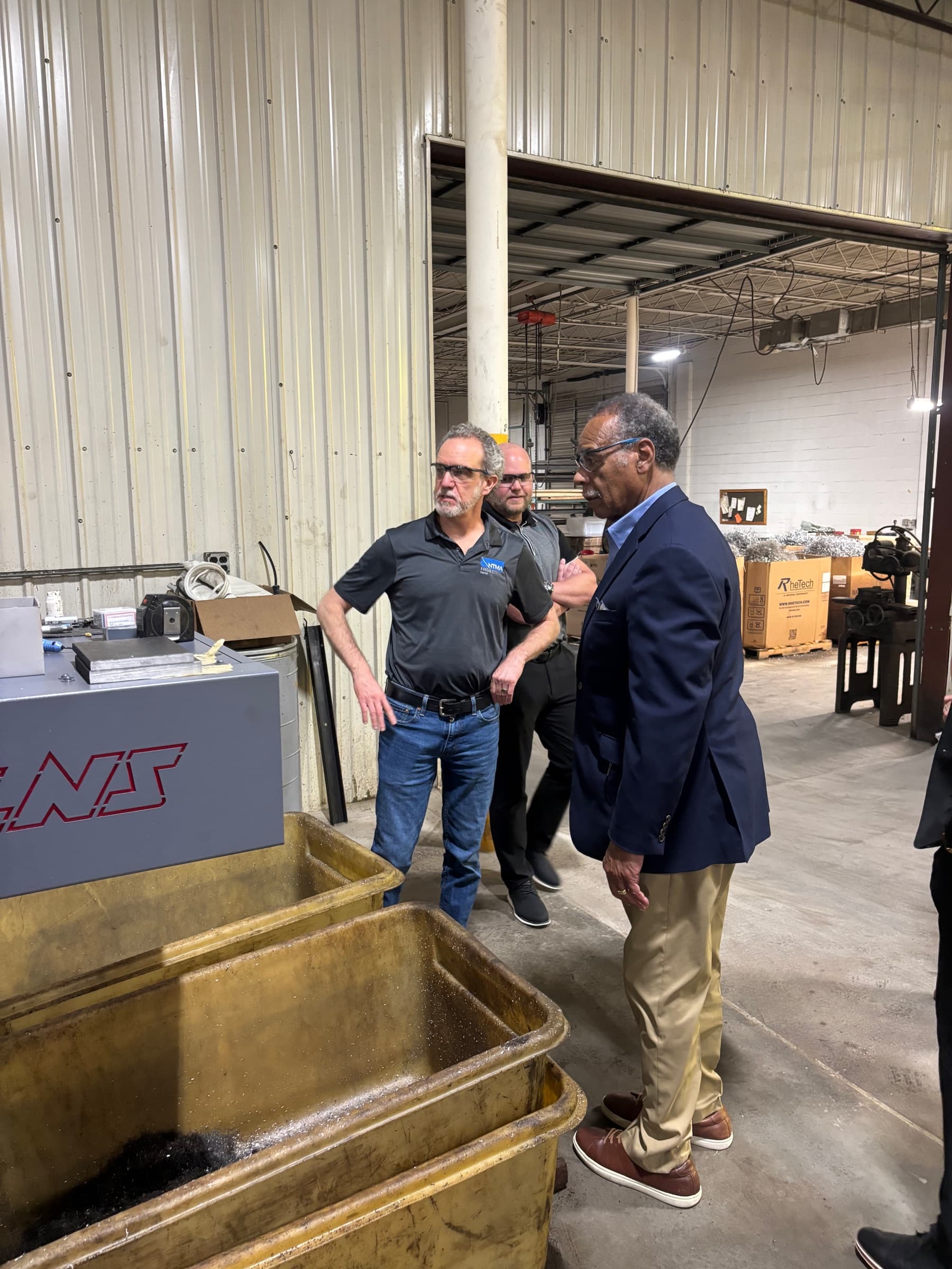 Congressman Emanuel Cleaver in a blue blazer looking over a machine with two shop staff