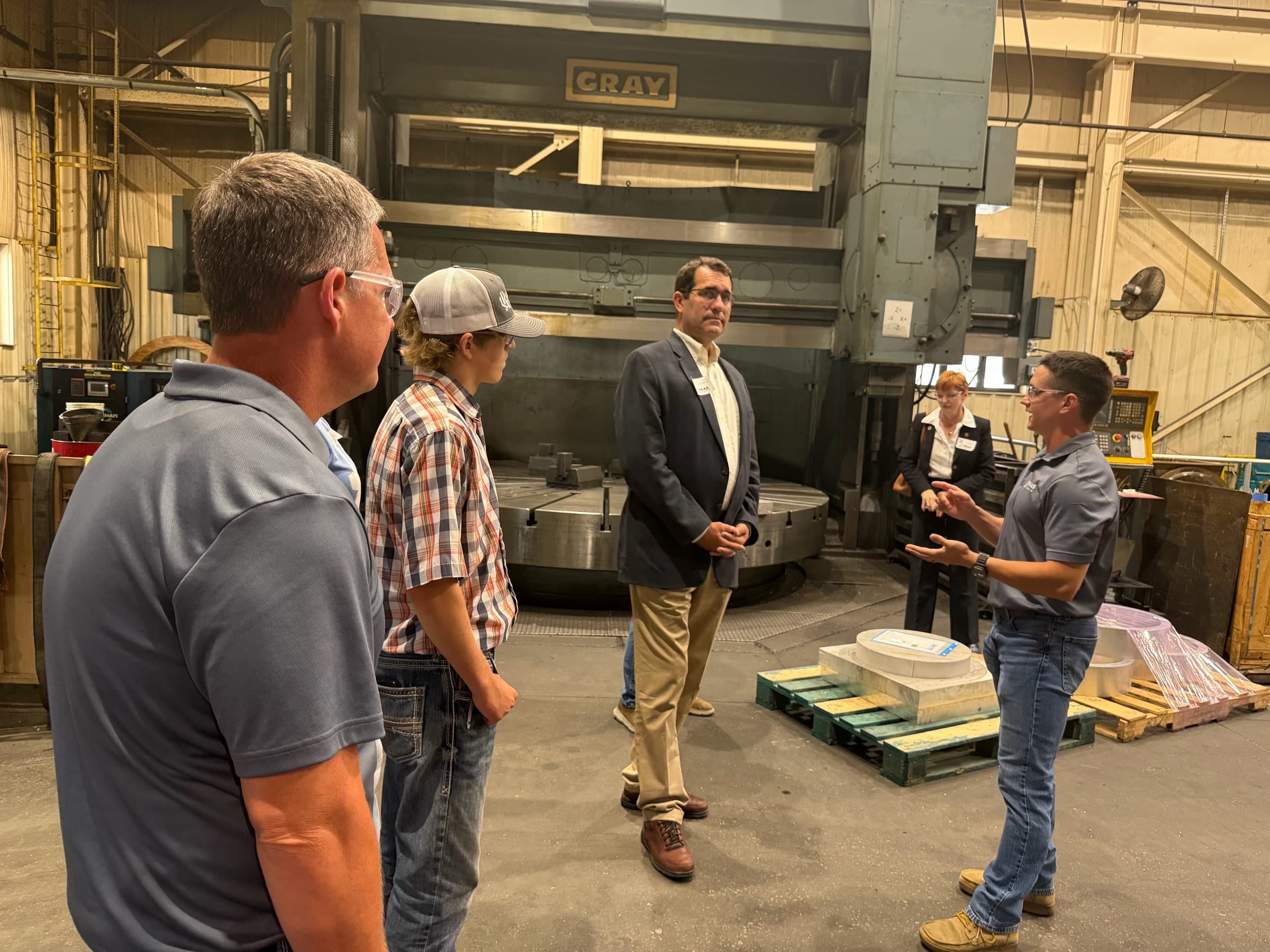 Congressman Derek Schmidt in a blazer listening to a shop owner on a foundry floor beside a large vertical boring mill
