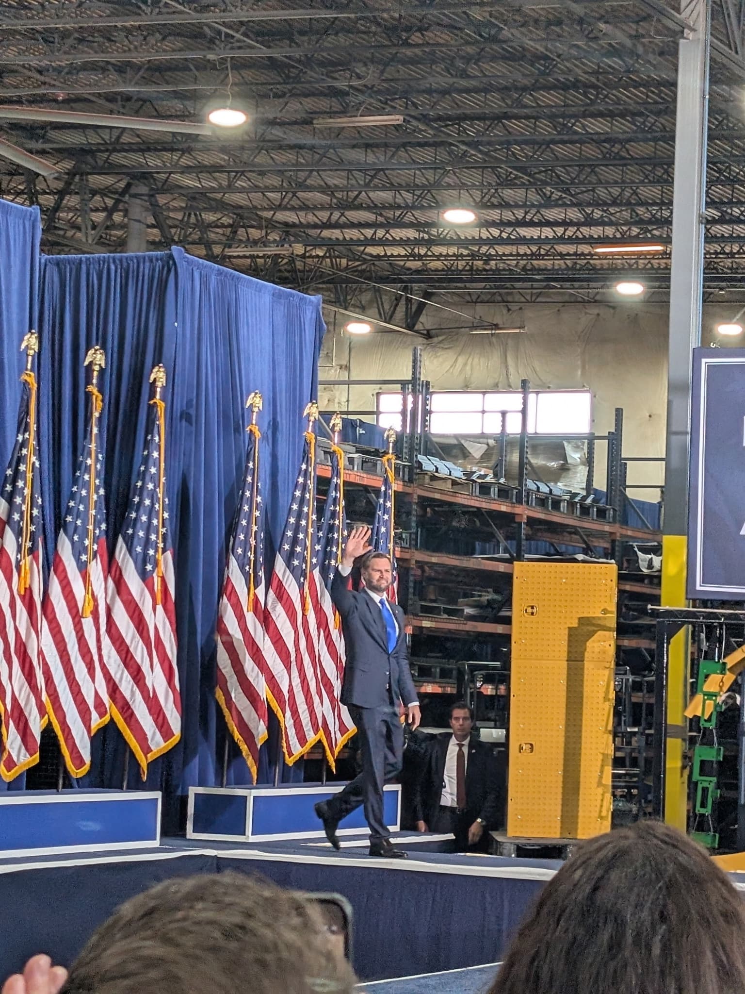 Vice President Vance waving as he walks on stage in front of a row of American flags inside a manufacturing plant