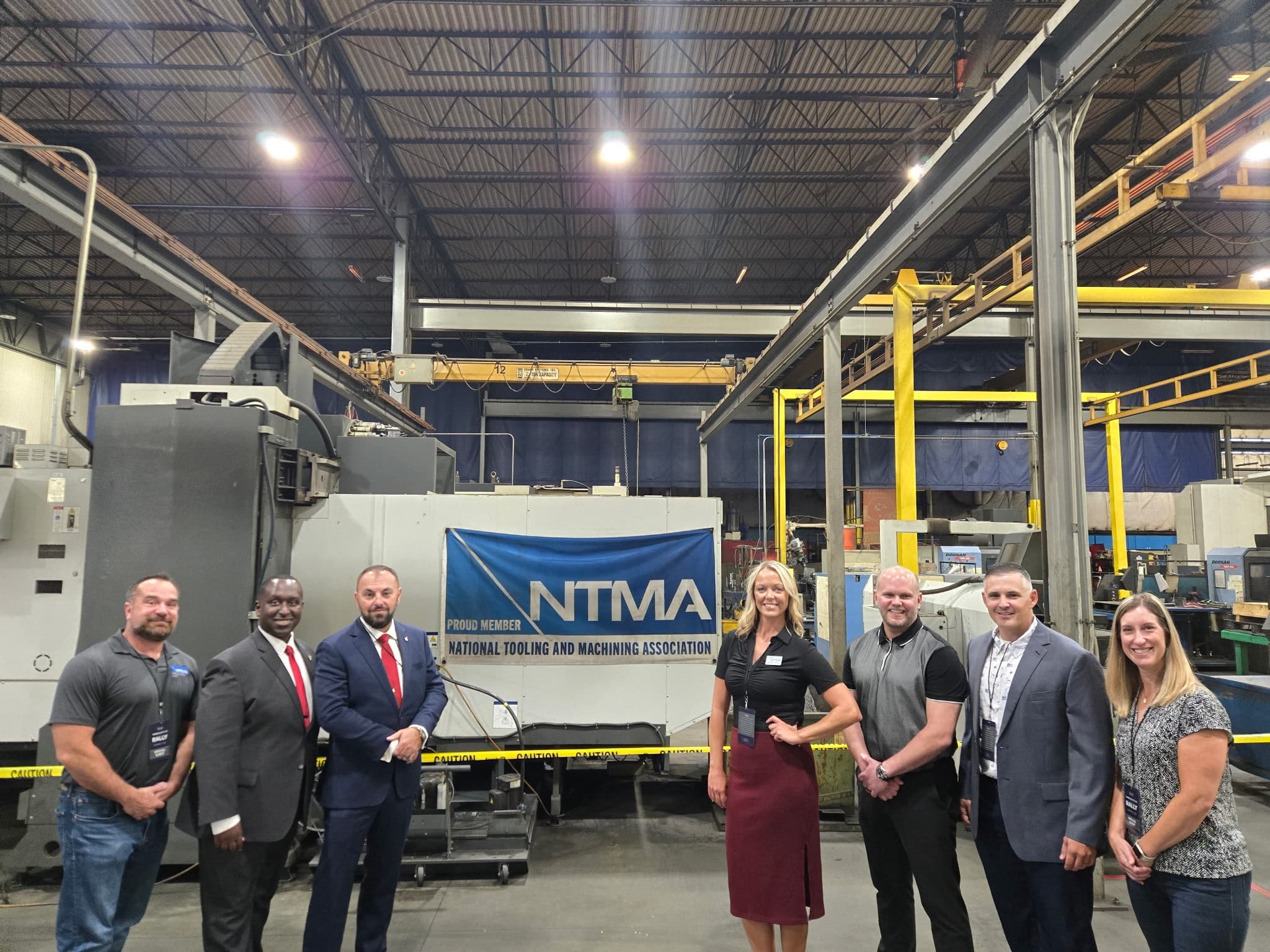 Eight people in front of a Proud Member NTMA banner on a plant floor, a CNC machine behind them
