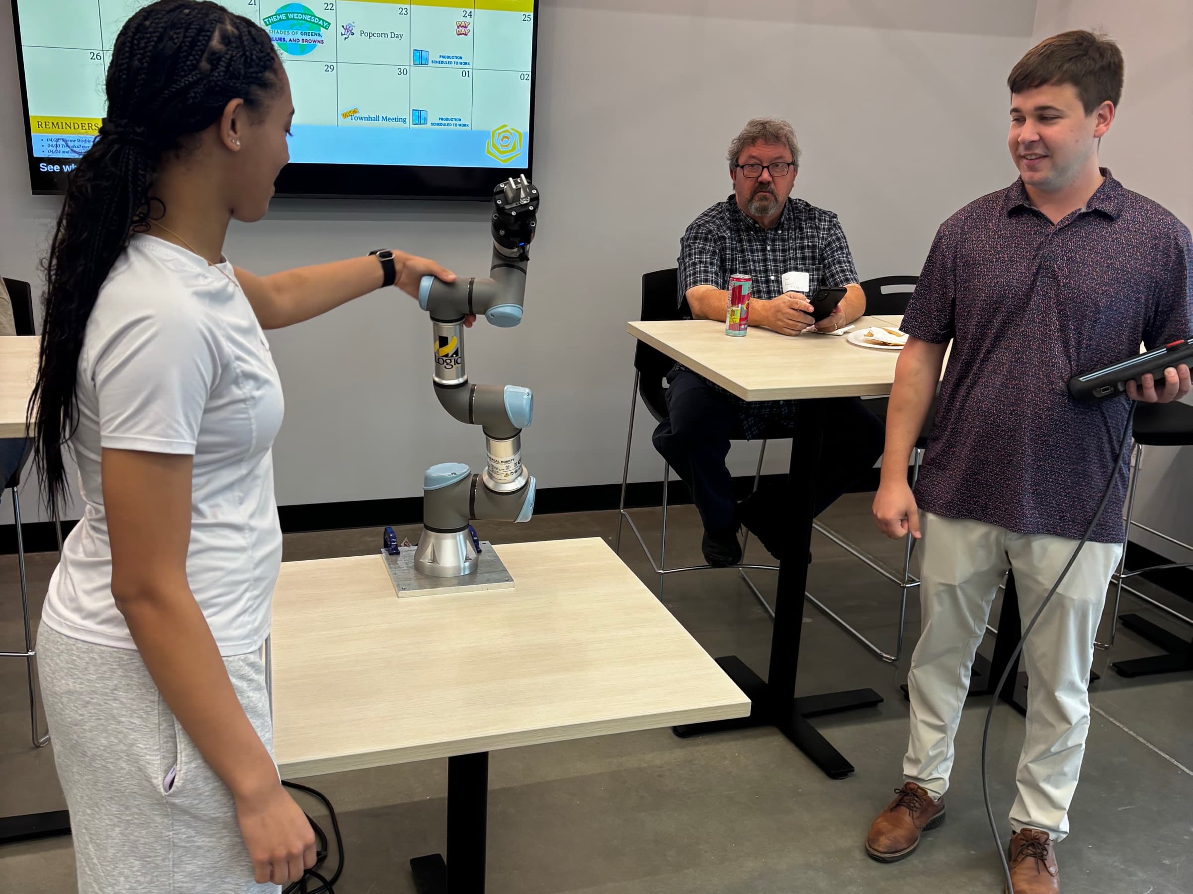 A young woman guiding a collaborative robot arm by hand while an instructor holding the teach pendant looks on