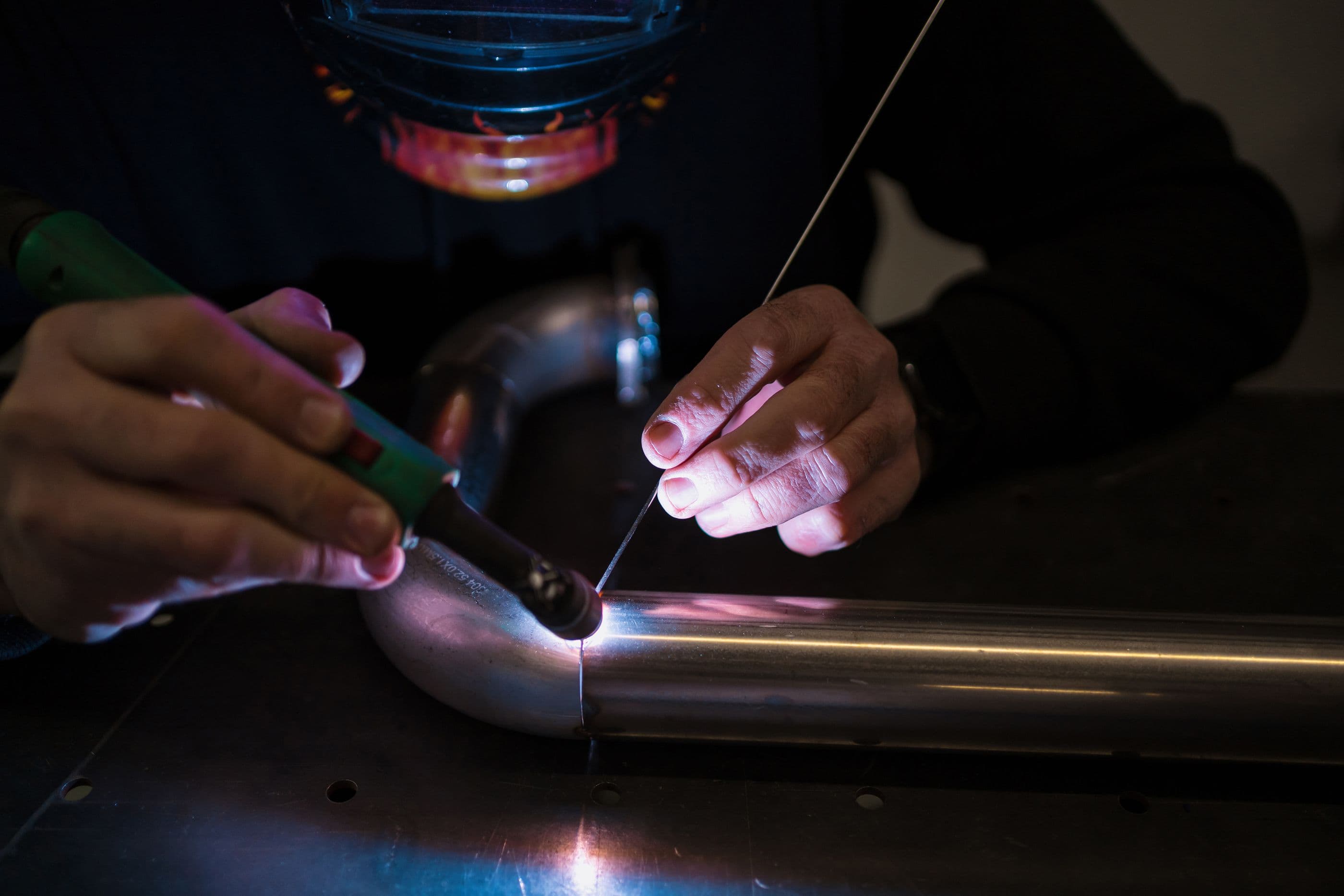 Hands guiding a TIG torch along a stainless pipe joint