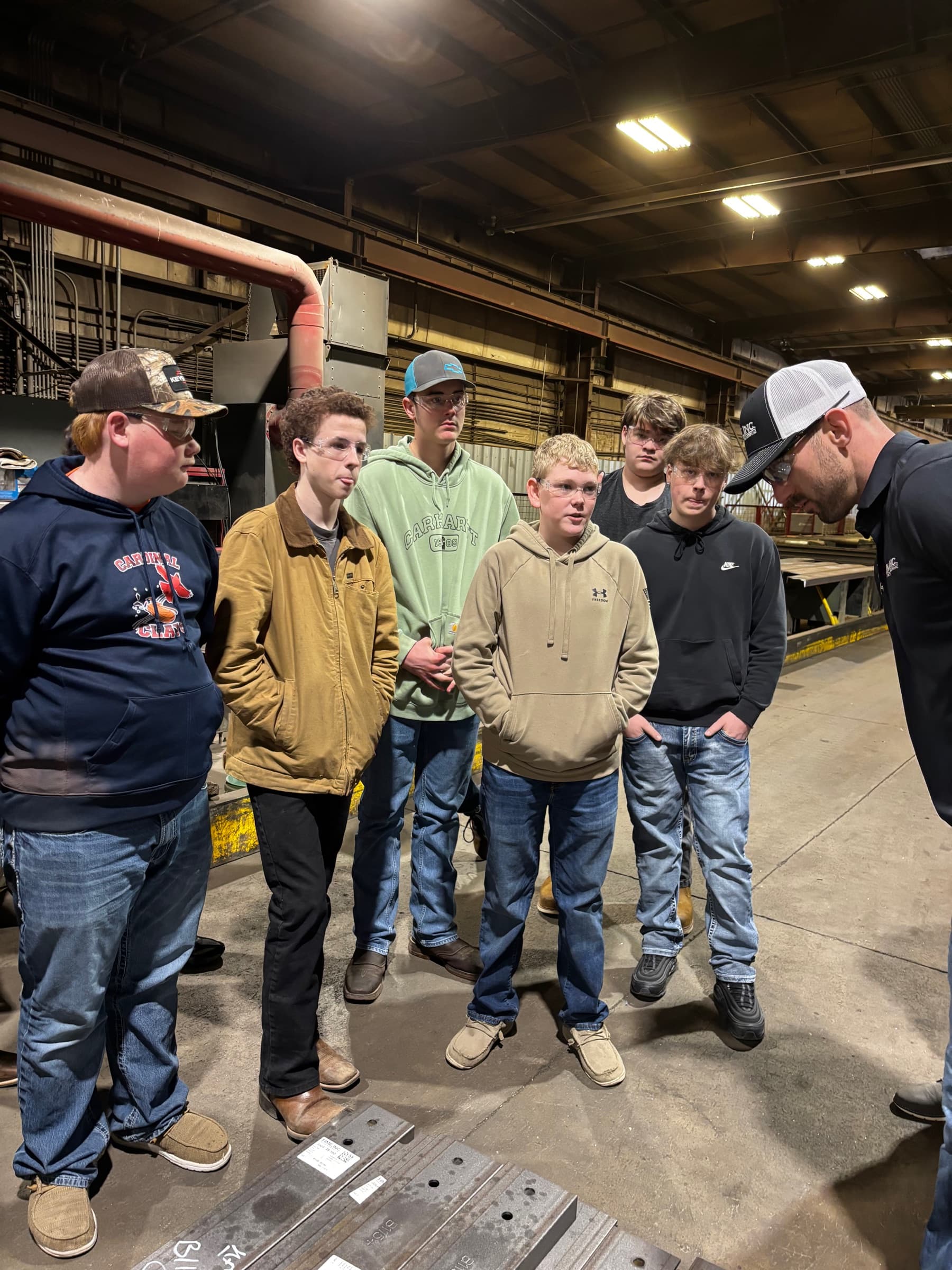 Six teenage boys listening closely to a shop guide in a cap on a plant floor