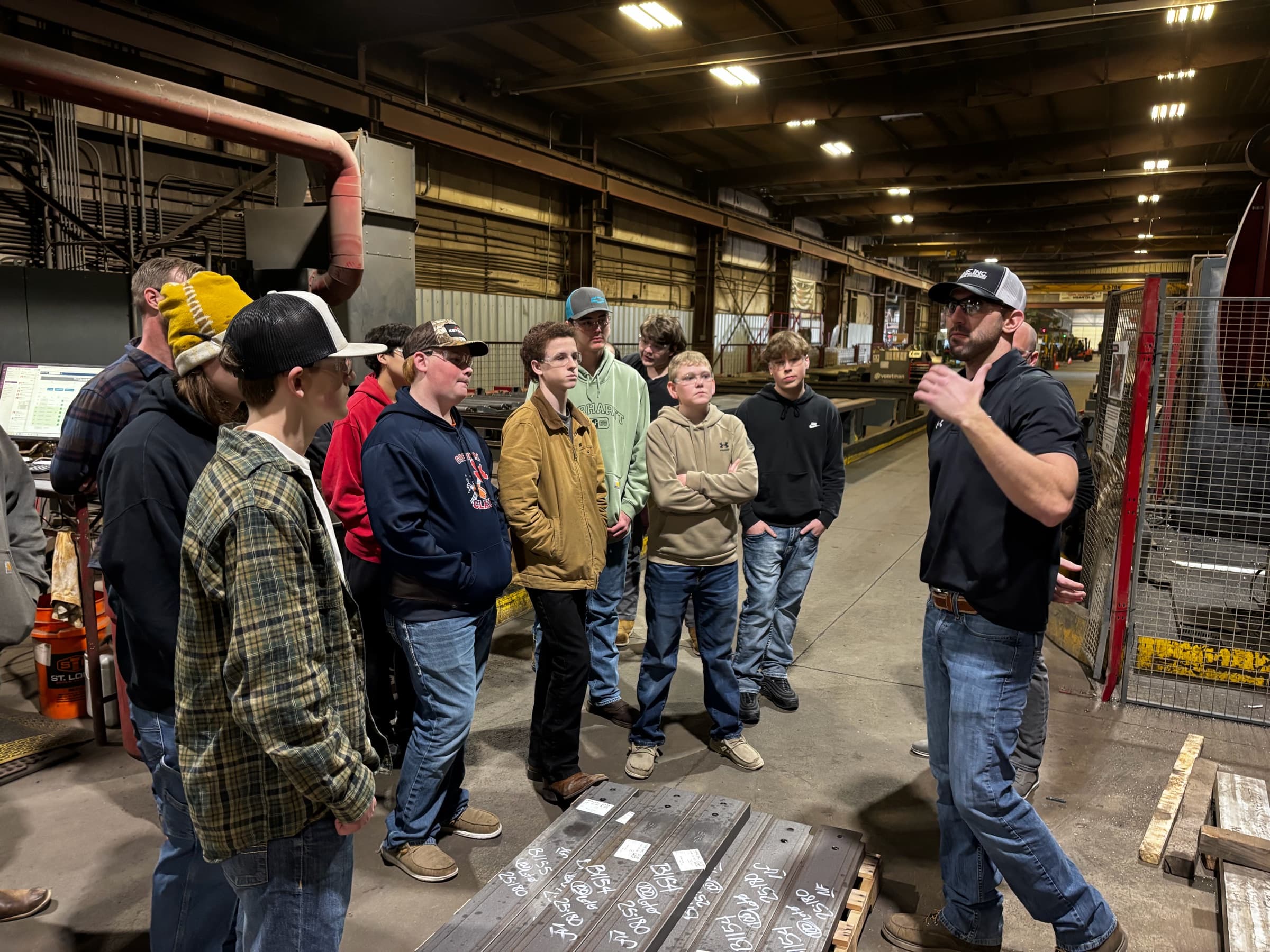 A dozen high school students in a fabrication plant listening to a guide beside a rack of steel plate