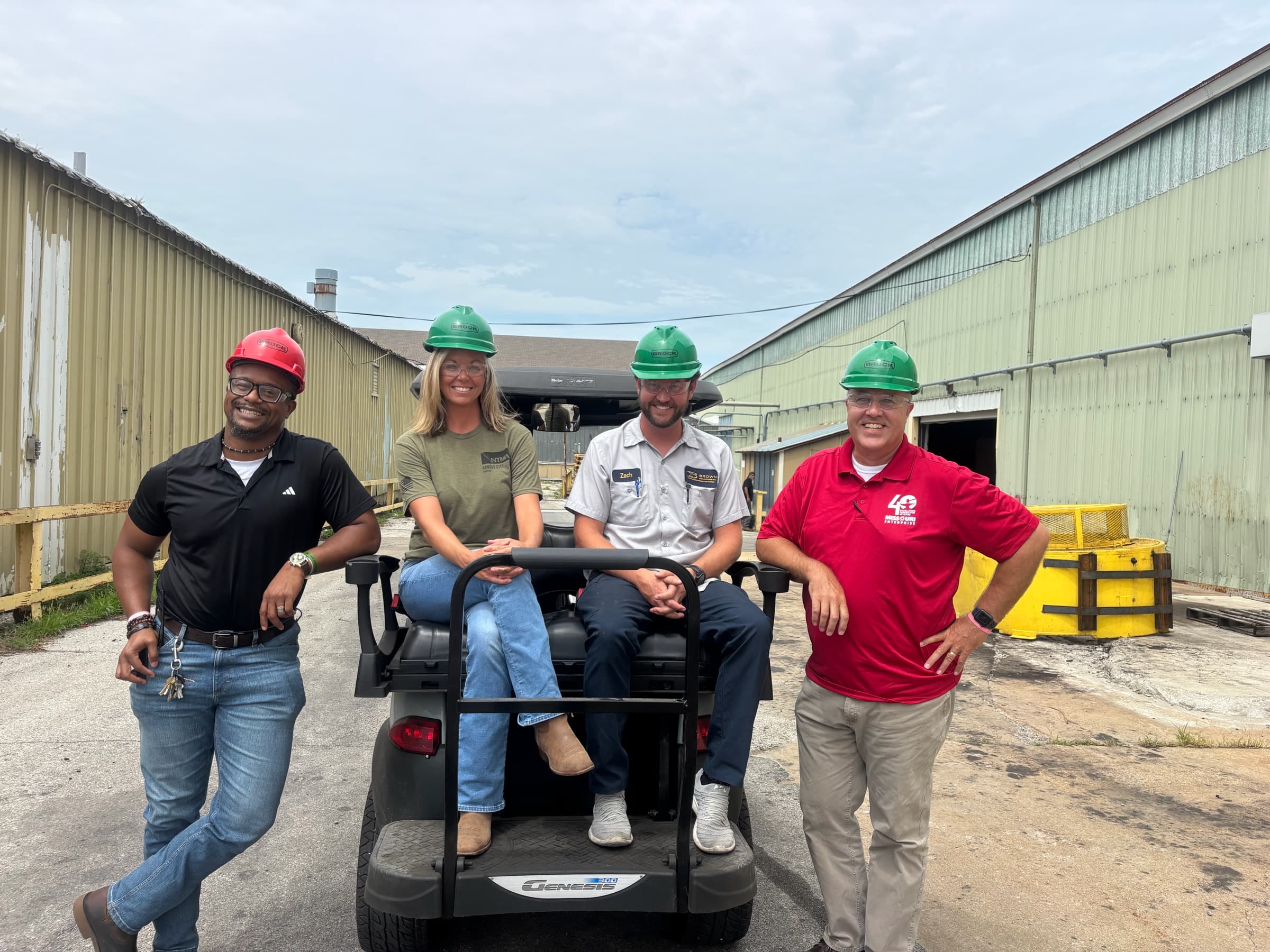 Four people in green and red hard hats around a golf cart between two plant buildings