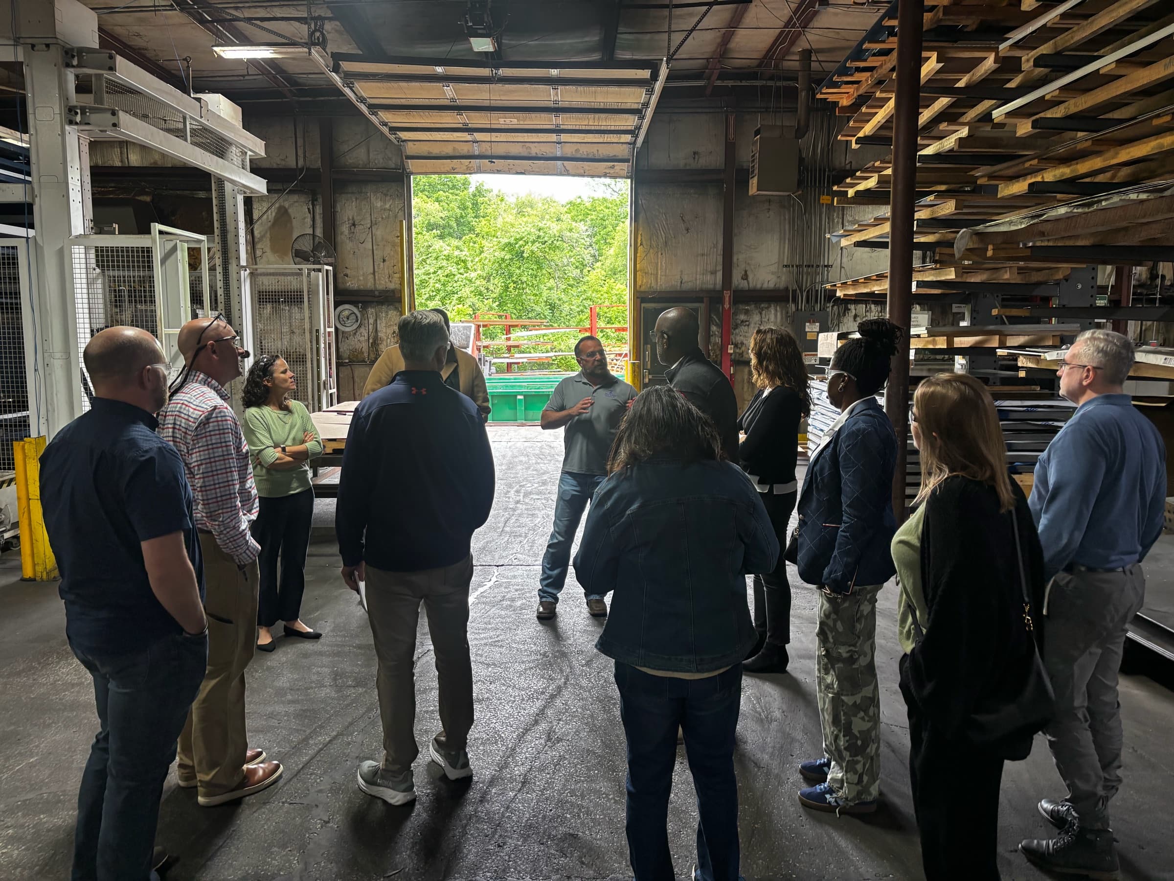 A tour group at a loading dock listening to a host, racks of lumber and steel behind them