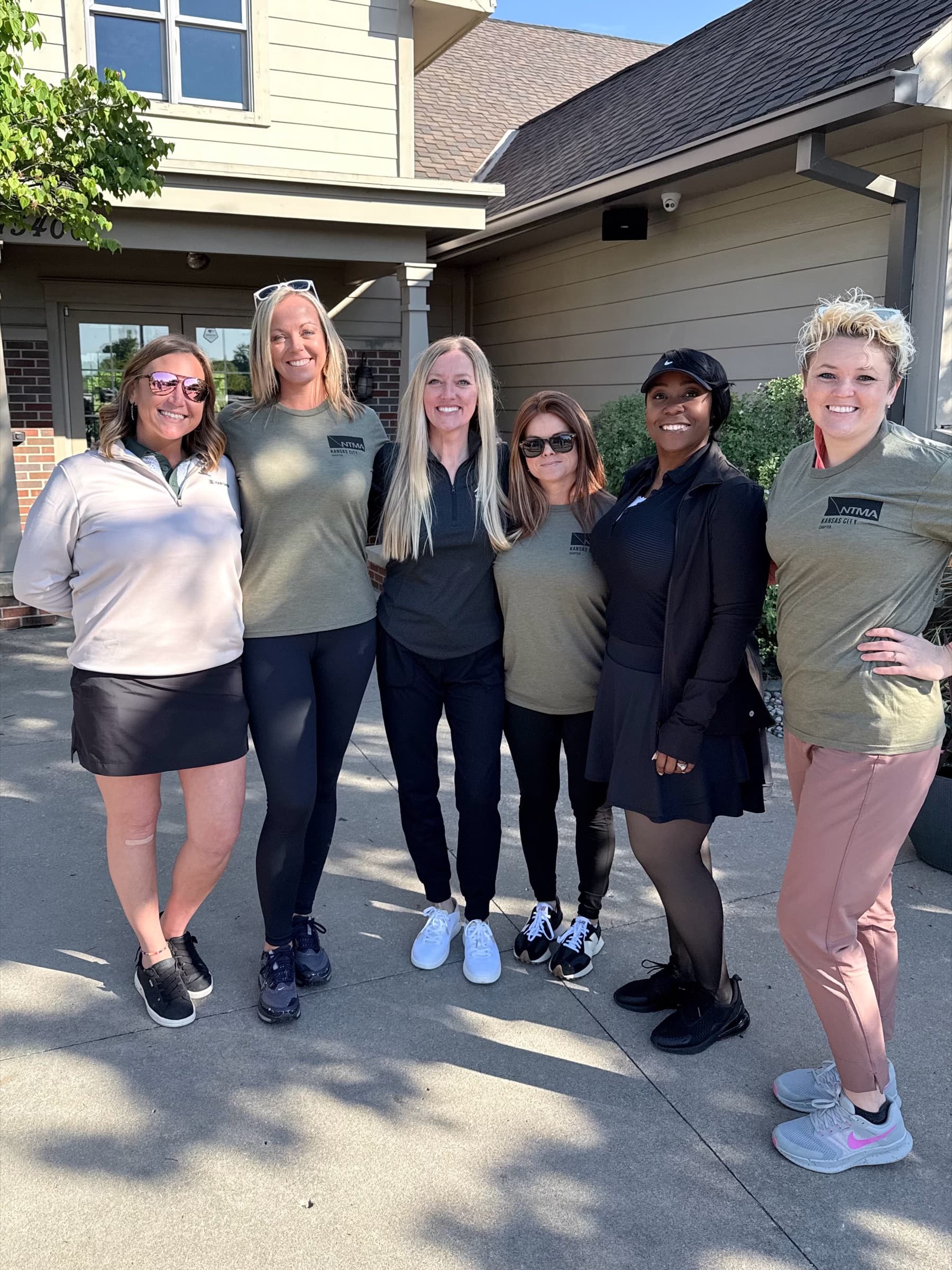 Six women in NTMA shirts outside a clubhouse on a sunny morning