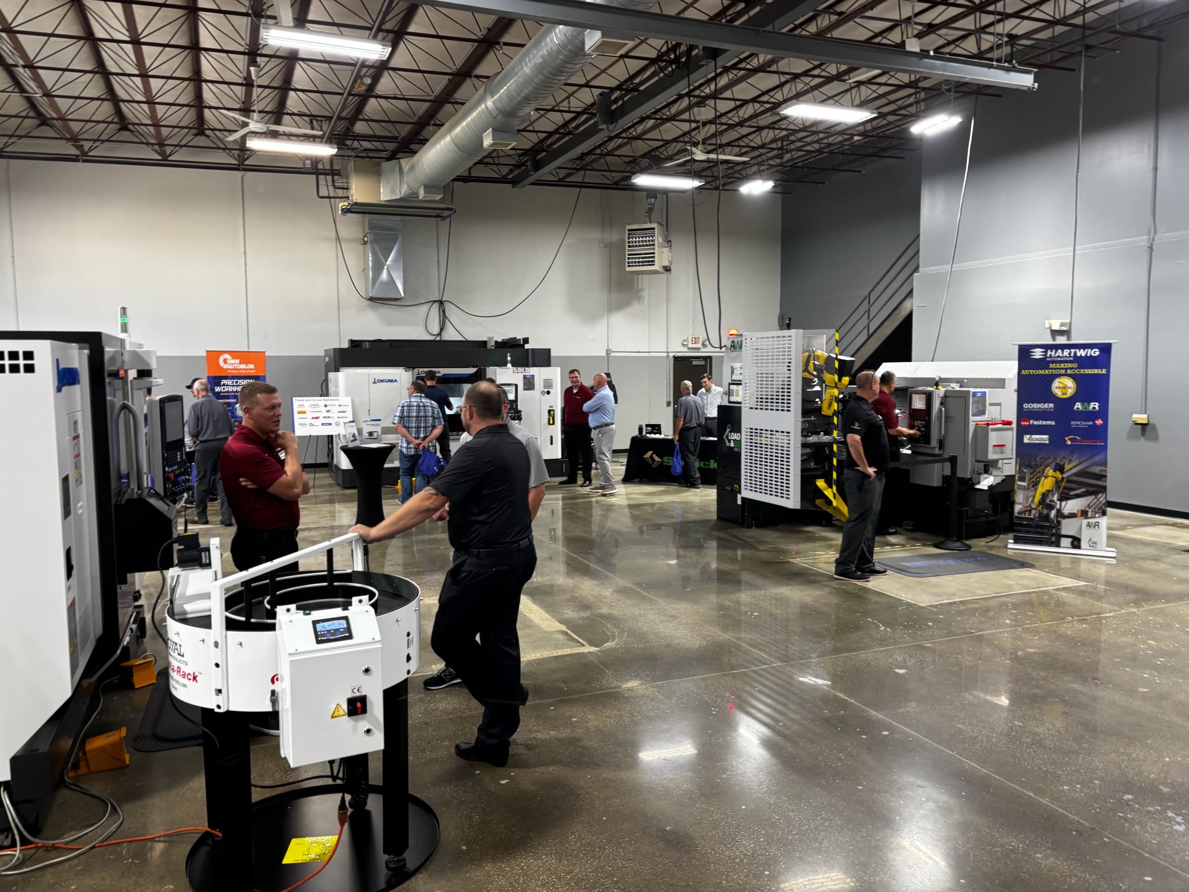 Members walking a machine tool showroom during an open house, a Hartwig banner and a CNC lathe at the back
