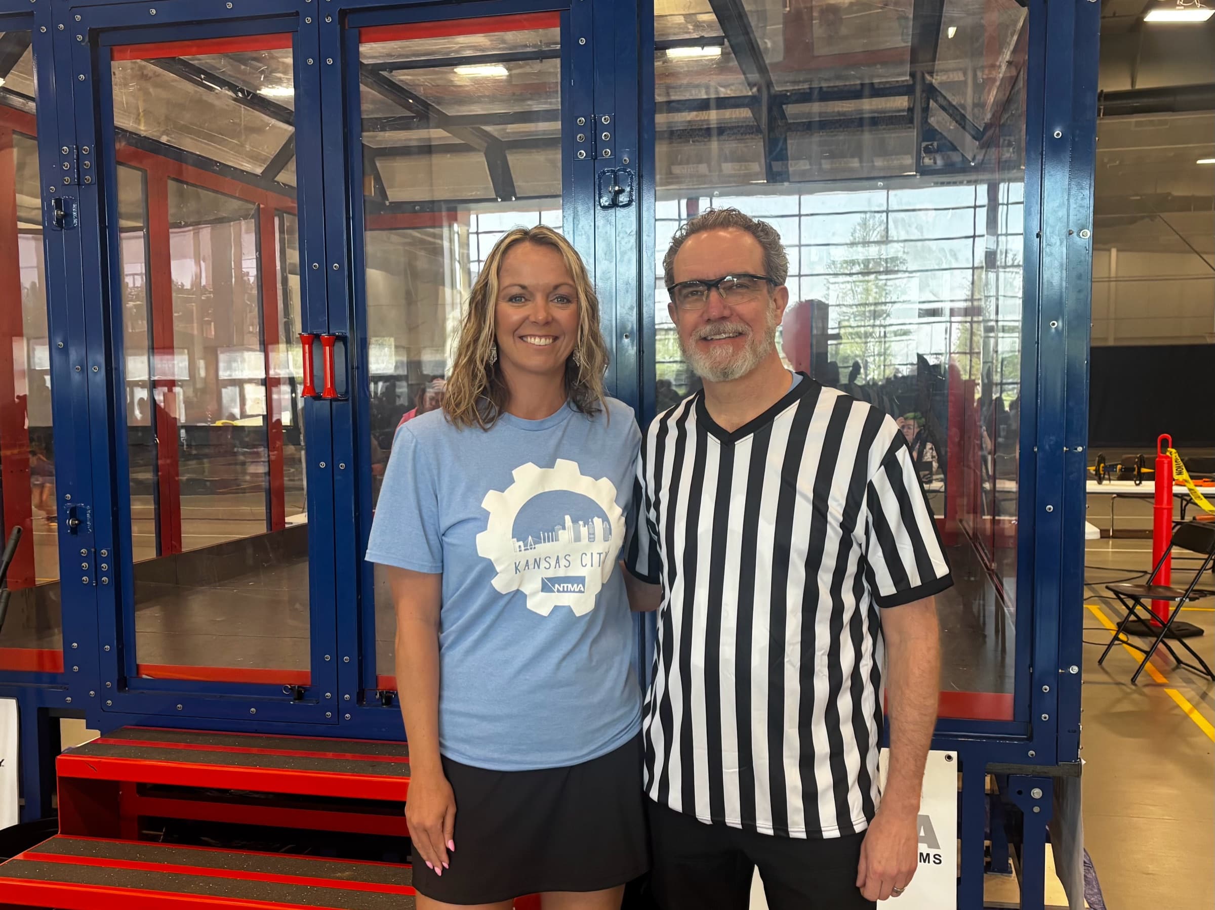 Katherine O'Toole in a Kansas City NTMA shirt beside a referee in front of the BotsKC robot arena