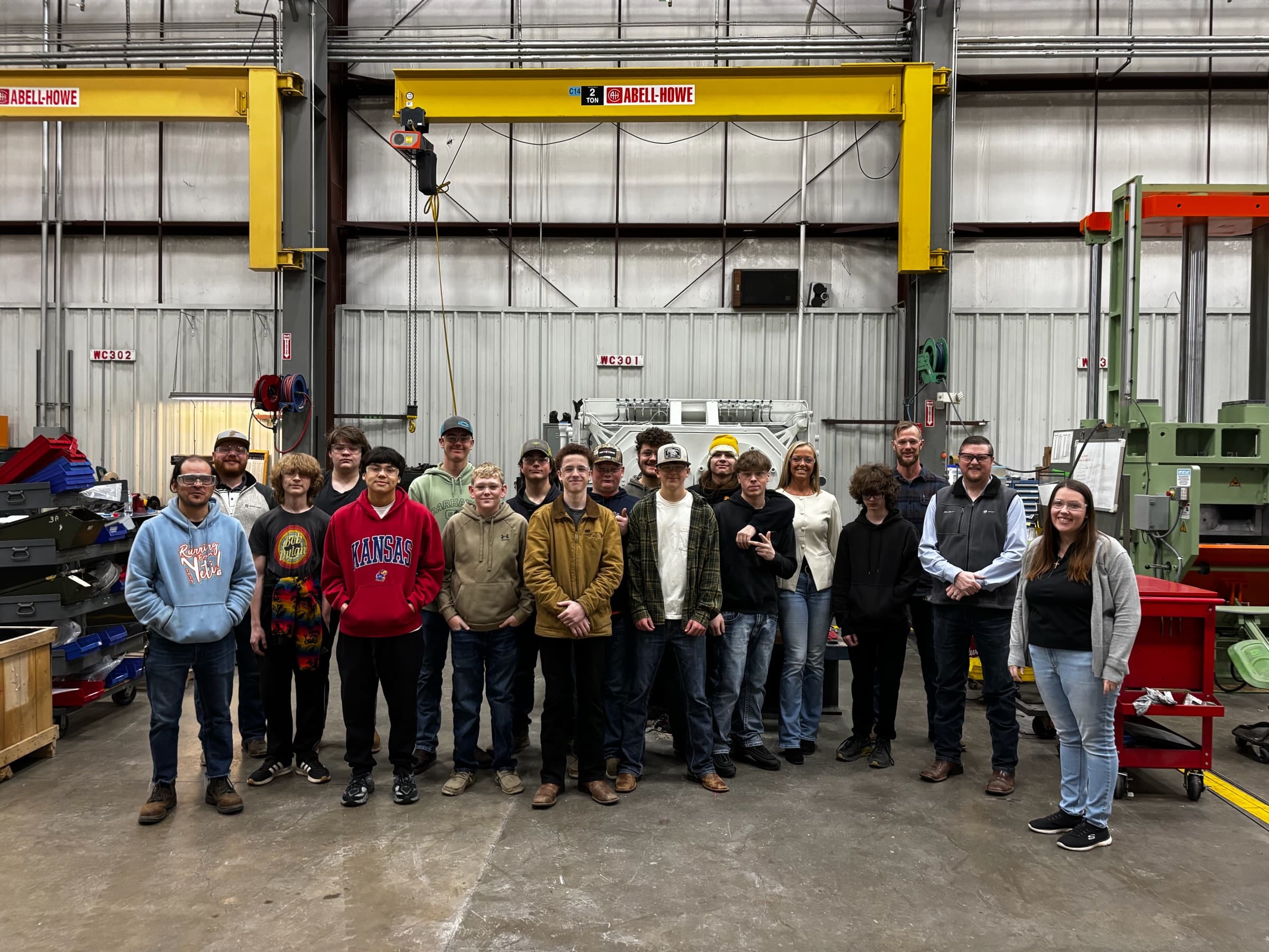 A class of high school students and their hosts on a plant floor beneath a yellow overhead crane