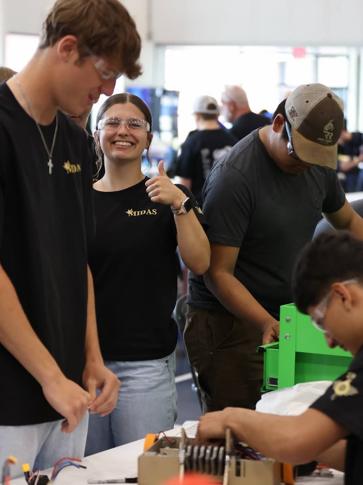 A student in safety glasses giving a thumbs up at a robotics table while classmates work on a build